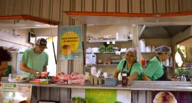 Foto de quatro pessoas, três dentro de um contêiner e uma fora. As pessoas dentro do food truck vestem verde, uma usa boné colorido e as outras duas usam toucas descartáveis. Ao fundo, várias prateleiras com alimentos e utensílios de cozinha. À frente, uma pessoa segura um pote com um pedaço de bolo. Há também um balcão com doces e talheres. Uma das pessoas do contêiner  sorri e coloca uma garrafa em cima do balcão. 