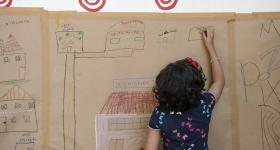 Foto de uma menina branca e de cabelos escuros até os ombros, que desenha com giz de cera em um papel kraft colado na parede. Ela está de costas e escreve em um retângulo desenhado ao lado de prédios e casas, ligados por ruas. No centro do desenho, há uma grande casa com telhado vermelho e paredes rosa claro. 