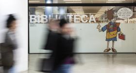 Foto da entrada da biblioteca da ECA. Sobre uma divisória de vidro, destaca-se em letras grandes e brancas a palavra ‘biblioteca’ e uma ilustração do cartunista Dorinho de uma pessoa de costas com uma mochila repleta de livros. Em primeiro plano e em desfoque, três pessoas caminham pelo saguão. 