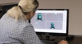 Foto de pessoa branca com cabelos loiros presos em um rabo de cavalo e vestindo uma camisa azul-clara de bolinhas brancas operando um computador com um mouse. Ela está sentada, de costas para a câmera, usando um software de modelagem 3D, que exibe um modelo tridimensional de um personagem ou objeto com aparência de boneco no monitor. 
