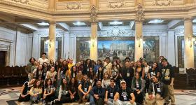 Foto de um grupo de cerca de 50 pessoas, majoritariamente mulheres jovens e brancas, todos com crachás pendurados no pescoço, dentro de um edifício antigo. O piso é feito de desenhos geométricos em mármore cinza, preto e laranja. Atrás das pessoas, colunas com luminárias e uma pintura grande pendurada na parede. O teto também é construído de abóbodas e desenhos esculpidos. 