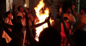 Foto de grupo de pessoas em um círculo em volta de uma fogueira. A silhueta das pessoas em primeiro plano é escura, enquanto as mais distantes, iluminadas pelo fogo têm tons avermelhados. O fundo é escuro.