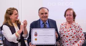 Foto de três pessoas em pé, lado a lado. No centro, um homem branco, idoso, vestindo terno escuro, camisa azul e gravata listrada, segurando e exibindo um certificado. À esquerda, uma mulher branca de cabelos loiros e longos, vestida com uma roupa preta e branca, está batendo palmas. À direita, há outra mulher branca, idosa, de cabelos castanhos curtos, vestindo uma blusa florida. Ao fundo, tela de projeção exibindo texto em azul e branco.