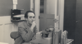 Foto em preto e branco de homem de cabelos curtos e escuros, usando paletó claro, sentado em uma cadeira atrás de uma mesa de escritório. Ele olha para a câmera enquanto leva a mão à boca. Sobre a mesa há uma máquina de escrever, uma maleta e uma bolsa de couro e um cinzeiro. Ao fundo, há portas de madeira com painéis de vidro e uma prateleira de parede com papéis. 