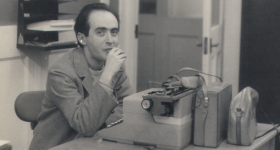 Foto em preto e branco de homem de cabelos curtos e escuros, usando paletó claro, sentado em uma cadeira atrás de uma mesa de escritório. Ele olha para a câmera enquanto leva a mão à boca. Sobre a mesa há uma máquina de escrever, uma maleta e uma bolsa de couro e um cinzeiro. Ao fundo, há portas de madeira com painéis de vidro e uma prateleira de parede com papéis. 