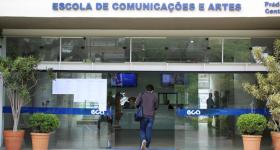 Foto da fachada de um prédio com portas de correr de vidro abertas. As portas têm uma faixa adesiva azul horizontal com o logotipo da Escola. Acima da entrada há um letreiro escrito Escola de Comunicações e Artes. A foto capta uma pessoa de costas entrando no prédio, que veste um agasalho escuro, uma calça jeans azul e está com uma bolsa pendurada no ombro esquerdo. Nas duas laterais da entrada há vasos de plantas ornamentais. No saguão interno há monitores de TV e pessoas ao fundo.