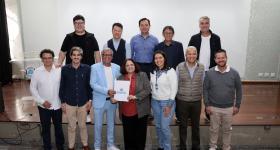 Foto de um grupo de 12 pessoas, de pé e posicionadas em duas fileiras. Sete pessoas à frente e cinco pessoas atrás, em cima de um palco de madeira, em sua maioria homens brancos. Ao centro, um homem negro, de cabelos brancos e curtos vestindo um terno azul claro e uma mulher parda, de cabelos de tamanho médio, vestindo um terno cinza e uma camisa vermelha, seguram e exibem uma pasta branca com a palavra “Suzano” impressa em azul. Ao fundo, uma tela branca e o piso de madeira.