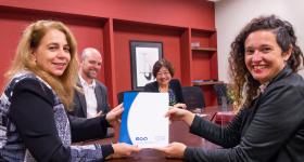 Foto de duas mulheres - ambas brancas, uma de cabelos longos e loiros e outra de cabelos castanhos cacheados - sorrindo enquanto posam segurando um documento com capa azul e branca, com os logotipos "eca" e "USP" em azul. Atrás delas, um homem branco careca e com barba e uma mulher amarela de cabelos castanhos e curtos também sorriem. Todos estão sentados em torno de uma mesa retangular de madeira. Ao fundo, uma parede vermelha com prateleiras embutidas e um quadro.