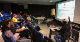 Foto de uma palestra em um auditório com cadeiras azuis, Uma plateia com cerca de 15 pessoas  assiste a uma apresentação expositiva de um homem sentado em uma mesa à  direita da foto e de costas. Ele usa fones de ouvido e boina branca, está sentado de frente para o público, apontando para uma tela grande onde um texto está projetado. Ele usa blusa preta e calça azul. O ambiente é pouco iluminado.