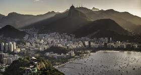 Foto da cidade do Rio de Janeiro a partir do alto. Paisagem litorânea com muitos prédios rodeados por montanhas, o mar repleto de barcos e iluminação do sol a partir do lado direito da imagem.