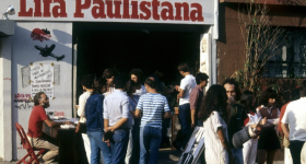 Foto da fachada do espaço cultural Lira Paulistana com uma multidão reunida na calçada. Pessoas conversam e folheiam livros, enquanto um homem sentado a uma mesa vermelha autografa obras. O letreiro “Lira Paulistana” está em destaque acima da entrada.