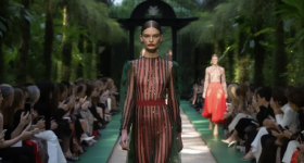 Mulher branca caminha por uma passarela cercada por vegetação verde em um desfile de moda. Ela veste um vestido longo verde e transparente com listras verticais em vermelho e preto, com detalhes brilhantes. Ela tem cabelos castanhos lisos que estão presos. Usa botas, cinto e batom vermelhos. Várias pessoas estão sentadas em ambos os lados da passarela, tirando fotos com celulares. No fundo desfocado, outra mulher branca se aproxima usando uma saia vermelha. 