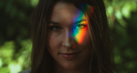 Foto de uma mulher de cabelos escuros e olhos claros. Sobre um de seus olhos, a projeção de um arco-íris. Ao fundo, folhagens desfocadas. 