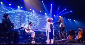 Foto de um palco durante um concerto. No centro, uma mulher negra, de cabelos crespos e loiros, está em pé diante de um microfone, usando um vestido escuro e um casaco branco e longo. Ao lado dela, um maestro de cabelos brancos usando um terno gesticula com uma das mãos erguidas, de frente para a orquestra. Atrás da cantora, um músico de roupas claras toca um violão sentado. À esquerda e à direita, membros da orquestra estão sentados com violinos, violoncelos e outros instrumentos.