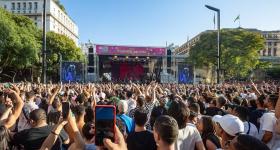 Foto de uma multidão de frente para um palco, no fundo da imagem, com uma placa rosa escrito Viaduto do Chá. Há um prédio baixo de cada lado do palco, assim como árvores dos dois lados. O céu está azul e sem nuvens.