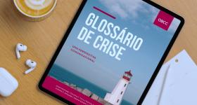 Foto de um leitor de livro digital preto sobre uma superfície de madeira de cor clara, tendo a seu lado uma xícara de café com a espuma da superfície decorada, fones sem fio auriculares brancos, carregador de fones branco, folhas de papel em branco e uma caneta de navegação branca. Na tela do leitor de livros está uma capa que tem como imagem de fundo a foto de um farol sobre rochas o mar e o horizonte azul com nuvens. O nome do livro está em fonte grande e branca, com complementos e destaques em vermelho 