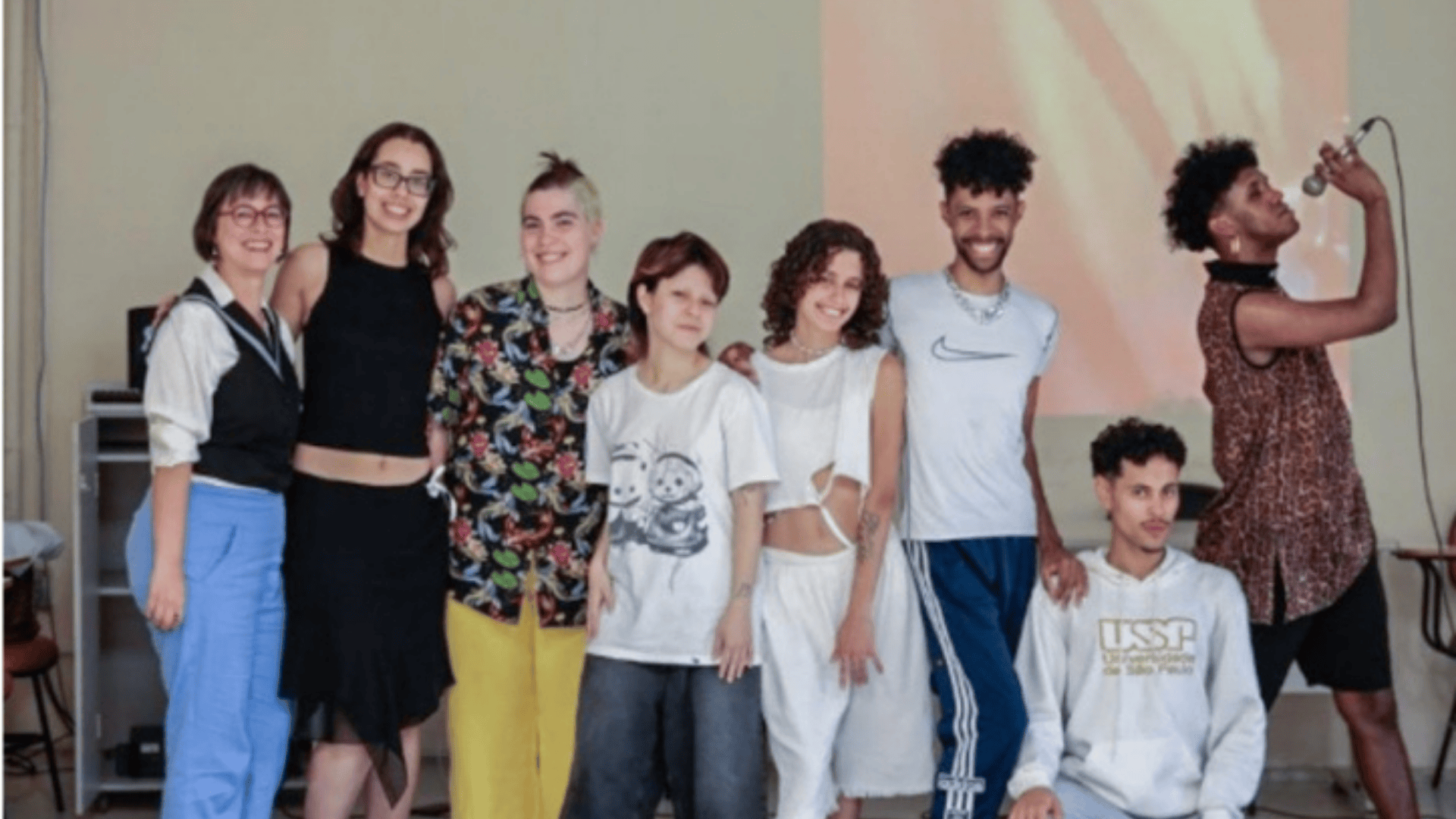 Foto de oito pessoas posando, de pé e sorrindo, em uma sala. Cinco das pessoas são brancas e três são rapazes negros, um deles está agachado e outro está de lado e segura um microfone em frente ao rosto. Todos usam roupas casuais como camisetas com alguma estampa e calças. O fundo tem paredes cinzas e uma projeção vermelha e amarela. 